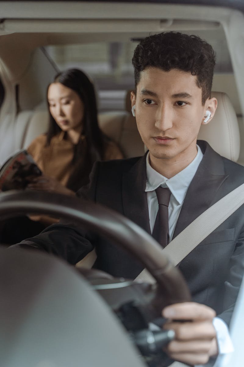 Professional driver in black suit focused on the road with a passenger in the backseat.